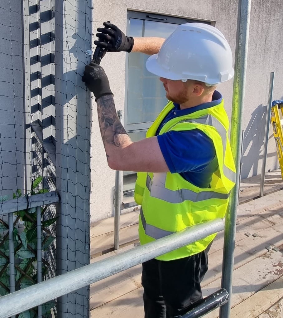 BW operative in hard hat and hi-vis fixing netting to a building facade from scaffolding
